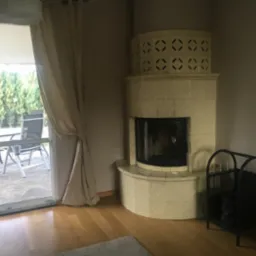 Jasny, kaflowy kominek z czarnym wkładem kominkowym w rogu pokoju z parkietem, obok czarny stojak na drewno, a w tle widok na taras i iglaki przez otwarte drzwi balkonowe.