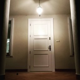 Artur Grela ELart Design - Białe trzyskrzydłowe drzwi wewnątrz budynku, oświetlone wiszącą lampą, widoczne kolumny i okna po bokach, ciemna podłoga.