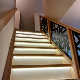 Artur Grela ELart Design - Nowoczesne schody z podświetlanymi stopniami i balustradą o geometrycznym wzorze z ciemnego metalu, drewniane elementy w jasnym odcieniu.