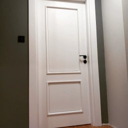 Artur Grela ELart Design - Białe drzwi wewnętrzne z czarną klamką, dzielone na dwie prostokątne sekcje z subtelnymi ramkami, zamontowane w szarej ścianie, widoczne drewniane panele podłogowe.