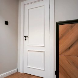 Artur Grela ELart Design - Białe drzwi z czarną klamką i ozdobnymi ramkami, widoczne na tle drewnianej podłogi i szarej ściany, obok fragmentu drewnianej komody.