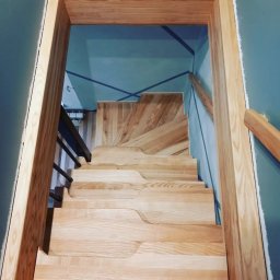 Artur Grela ELart Design - Widok z góry na jasne, drewniane schody z nieregularnymi krawędziami stopni, prowadzące w górę, z metalową balustradą po lewej stronie i drewnianą poręczą po prawej, na tle błękitnych ścian...