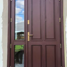 Brązowe dwuskrzydłowe drzwi wejściowe z szybą, widoczne ślady prac wykończeniowych na ścianie, odbicie osoby w odblaskowej kamizelce w szybie.