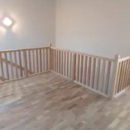 Jasna drewniana balustrada z pionowymi szczeblami i zaokrąglonymi słupkami na górnym poziomie domu, widok z góry na parkietową podłogę.