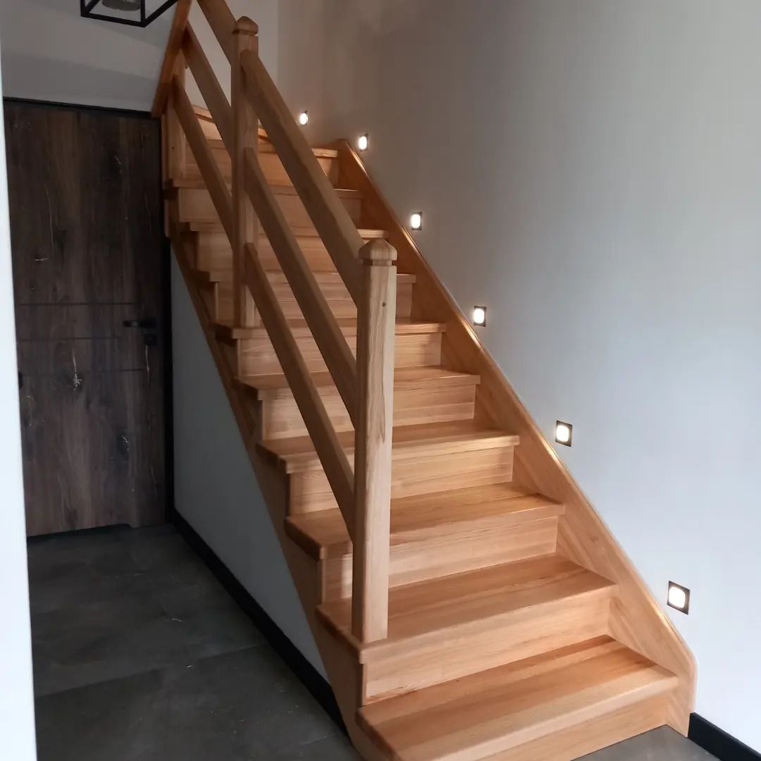 Jasne, drewniane schody z balustradą i oświetleniem punktowym wzdłuż ściany, widok od dołu, obok ciemne drzwi.
