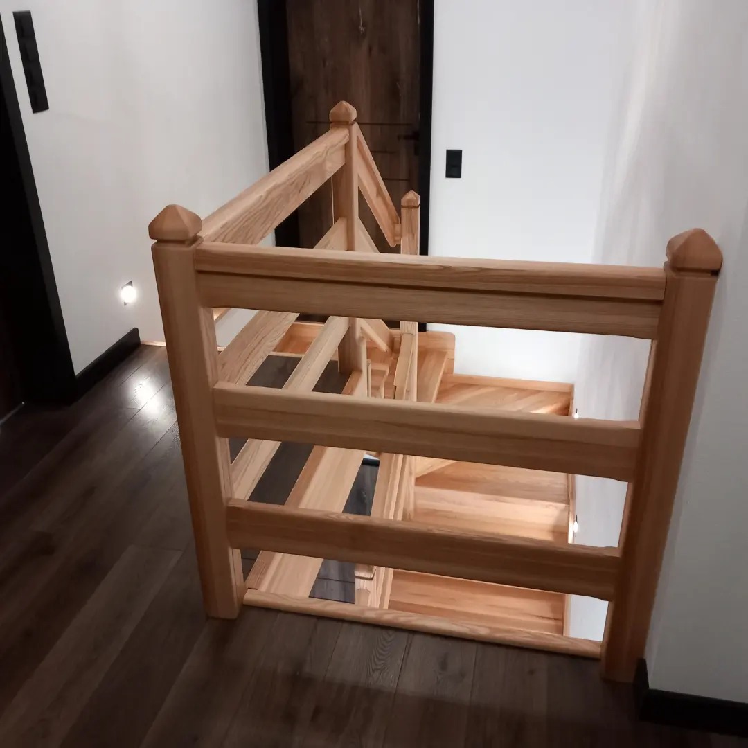 Drewniane schody z jasnego drewna z balustradą, widok z góry na dół, ciemna podłoga i drzwi w tle.