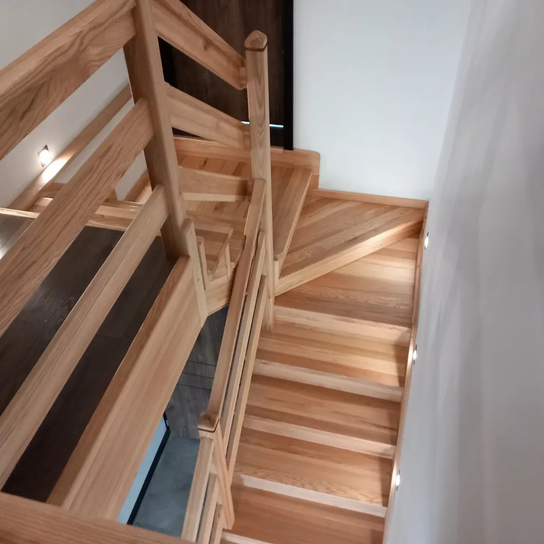 Jasne, drewniane schody z balustradą, widok z góry, oświetlenie punktowe w ścianie.