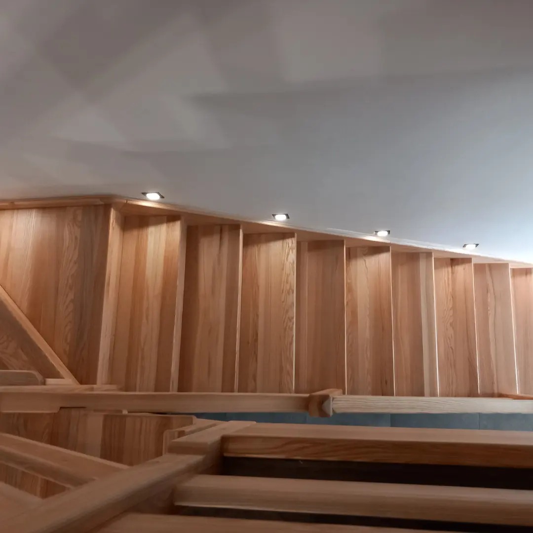 Jasne, drewniane schody z oświetleniem punktowym w suficie, widok z dołu, ukazujący konstrukcję i poręcz.