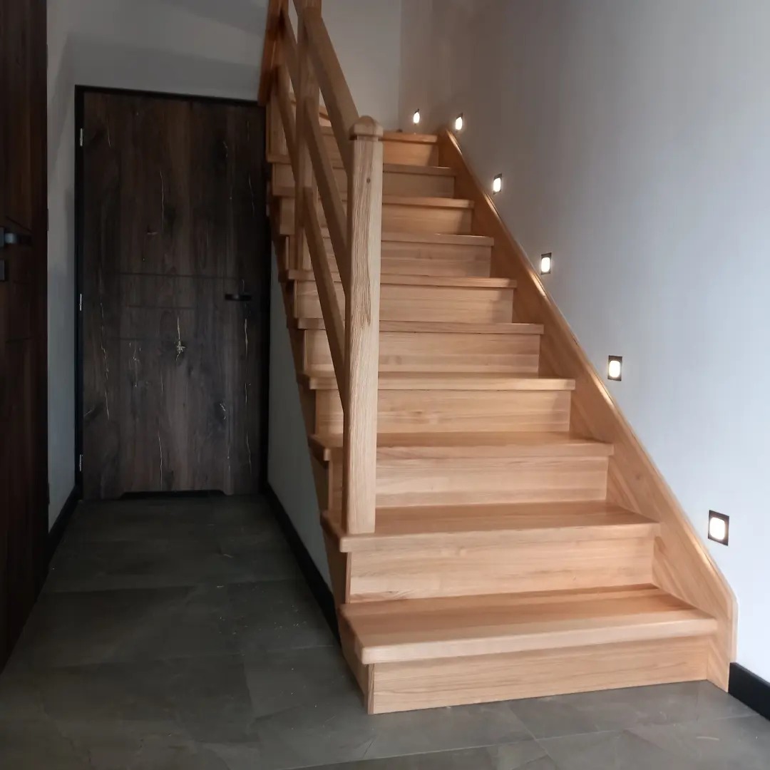 Jasne, drewniane schody ze ścianą oświetloną punktowymi lampkami, widok z dołu na tle ciemnych drzwi.