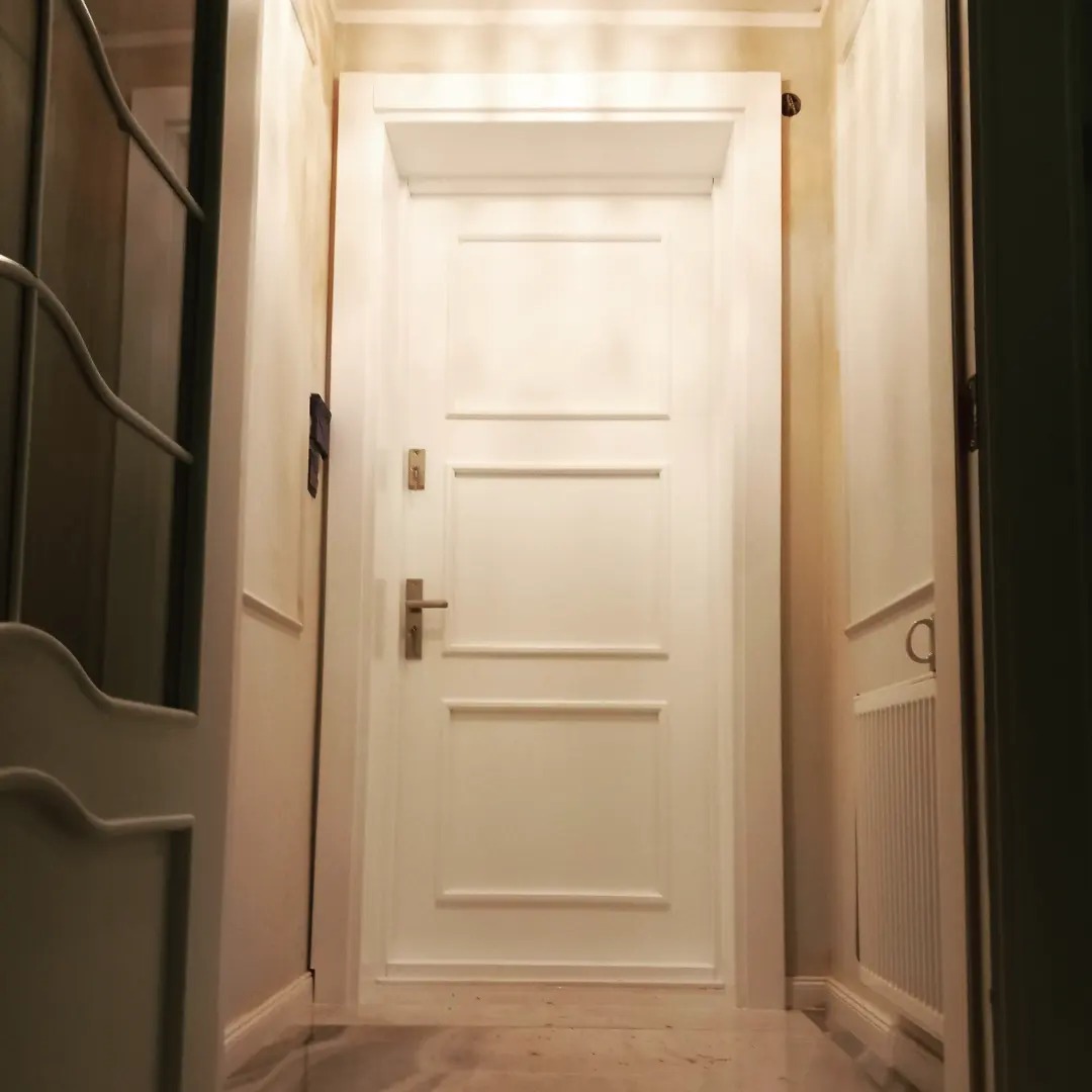 Białe drzwi wewnętrzne z trzema prostokątnymi panelami, widoczne z perspektywy korytarza z marmurową podłogą i dekoracyjnymi listwami ściennymi.