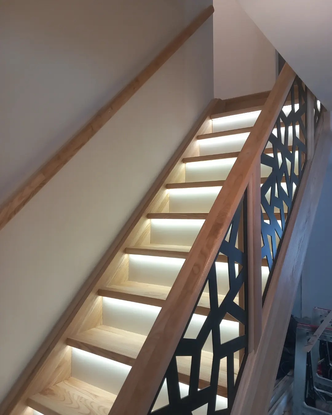 Drewniane schody z podświetlanymi stopniami i nowoczesną, geometryczną balustradą w kolorze czarnym, wykonane z jasnego drewna.