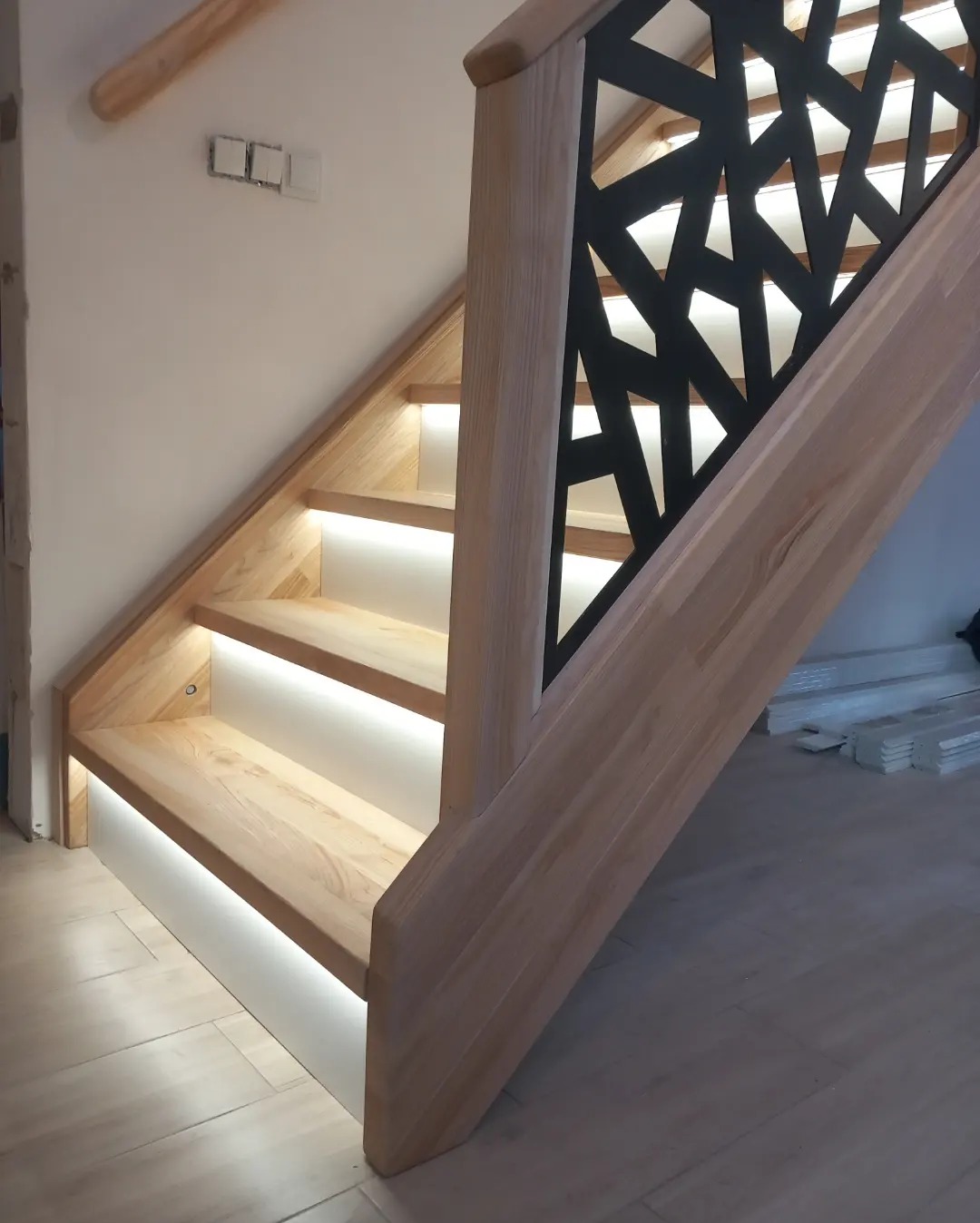 Nowoczesne schody z jasnego drewna z podświetleniem LED pod stopniami i czarną, geometryczną balustradą, widok z dołu.