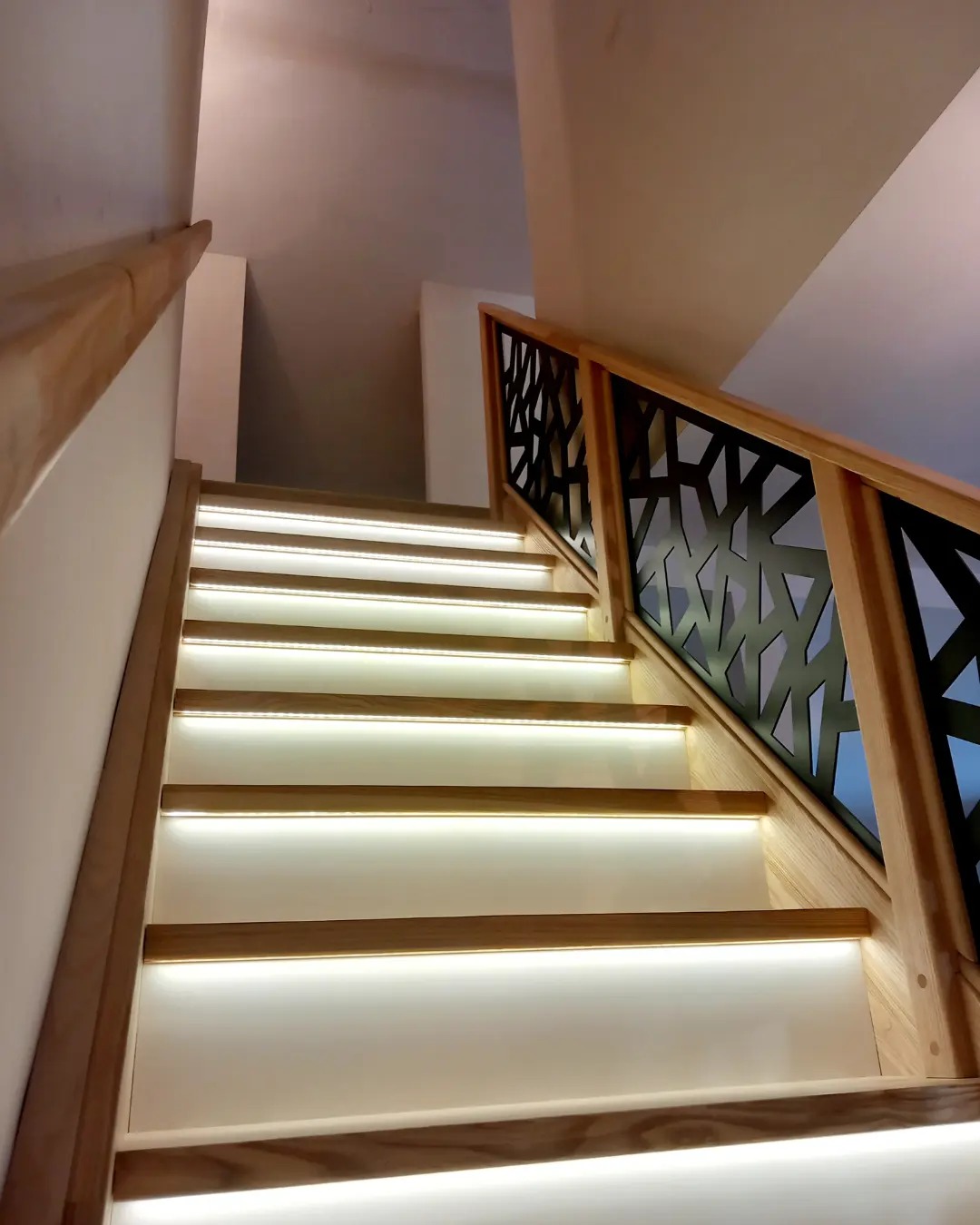 Nowoczesne schody z podświetlanymi stopniami i balustradą o geometrycznym wzorze z ciemnego metalu, drewniane elementy w jasnym odcieniu.