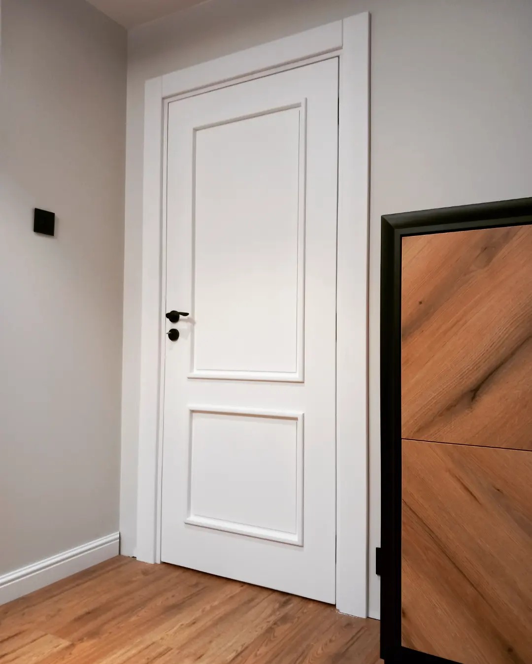 Białe drzwi z czarną klamką i ozdobnymi ramkami, widoczne na tle drewnianej podłogi i szarej ściany, obok fragmentu drewnianej komody.