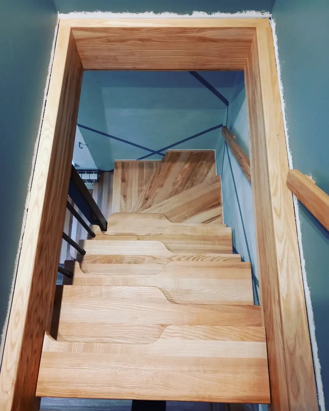 Widok z góry na jasne, drewniane schody z nieregularnymi krawędziami stopni, prowadzące w górę, z metalową balustradą po lewej stronie i drewnianą poręczą po prawej, na tle błękitnych ścian...