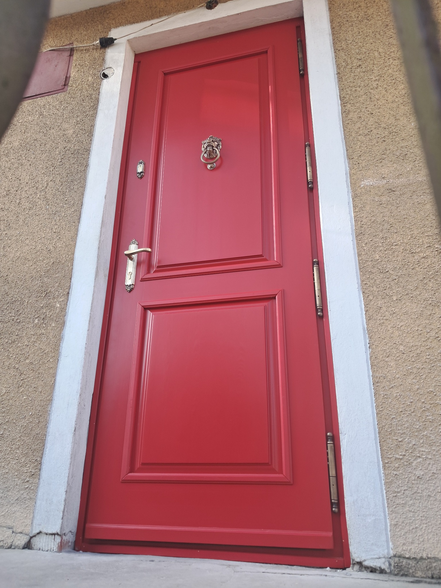 Czerwone drzwi wejściowe z ozdobną kołatką w kształcie lwa i złotą klamką, osadzone w białej futrynie na tle beżowej ściany o chropowatej fakturze.