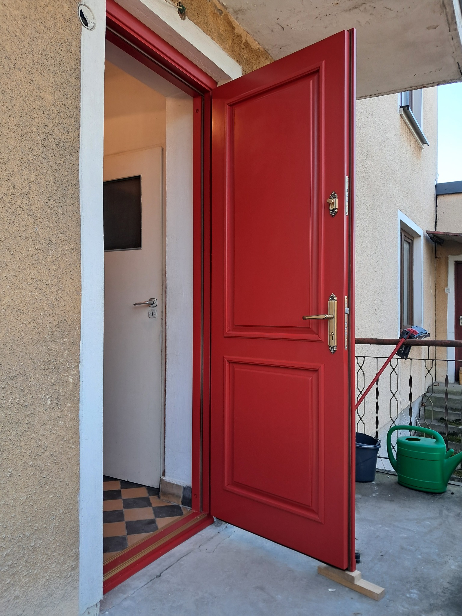 Czerwone drzwi wejściowe otwarte na zewnątrz, widoczny fragment wnętrza z białą ścianą i podłogą w szachownicę, na zewnątrz balkon z narzędziami i konewką.