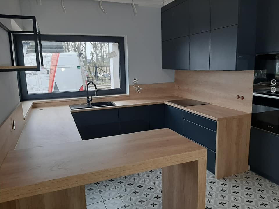 Nowoczesna kuchnia z ciemnymi szafkami i blatem imitującym drewno, wyspa kuchenna z blatem z widocznym usłojeniem, wzorzysta podłoga, okno z widokiem na samochód dostawczy.