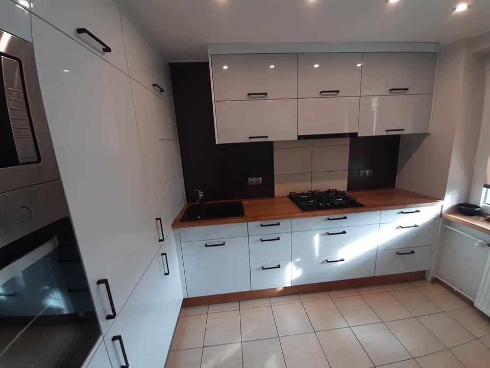 Nowoczesna kuchnia z białymi, lakierowanymi frontami, drewnianym blatem i czarnymi, minimalistycznymi uchwytami, widok na zabudowę piekarnika i płytę gazową.