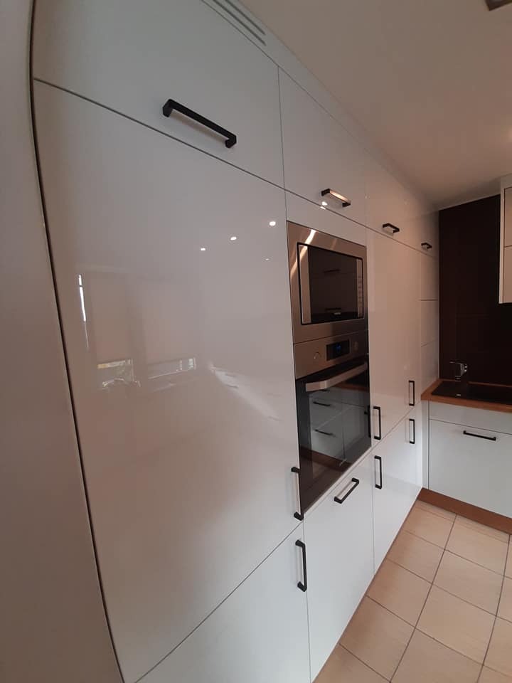 Białe, lakierowane fronty szafek kuchennych z czarnymi, prostokątnymi uchwytami, zabudowany piekarnik i mikrofalówka, jasna podłoga z płytek.