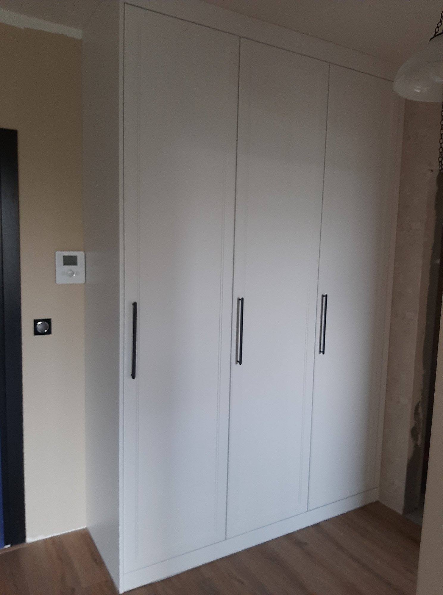 Biała, czterodrzwiowa szafa na wymiar z długimi, czarnymi uchwytami, zamontowana w mieszkaniu w Bydgoszczy, widoczny termostat na ścianie obok.