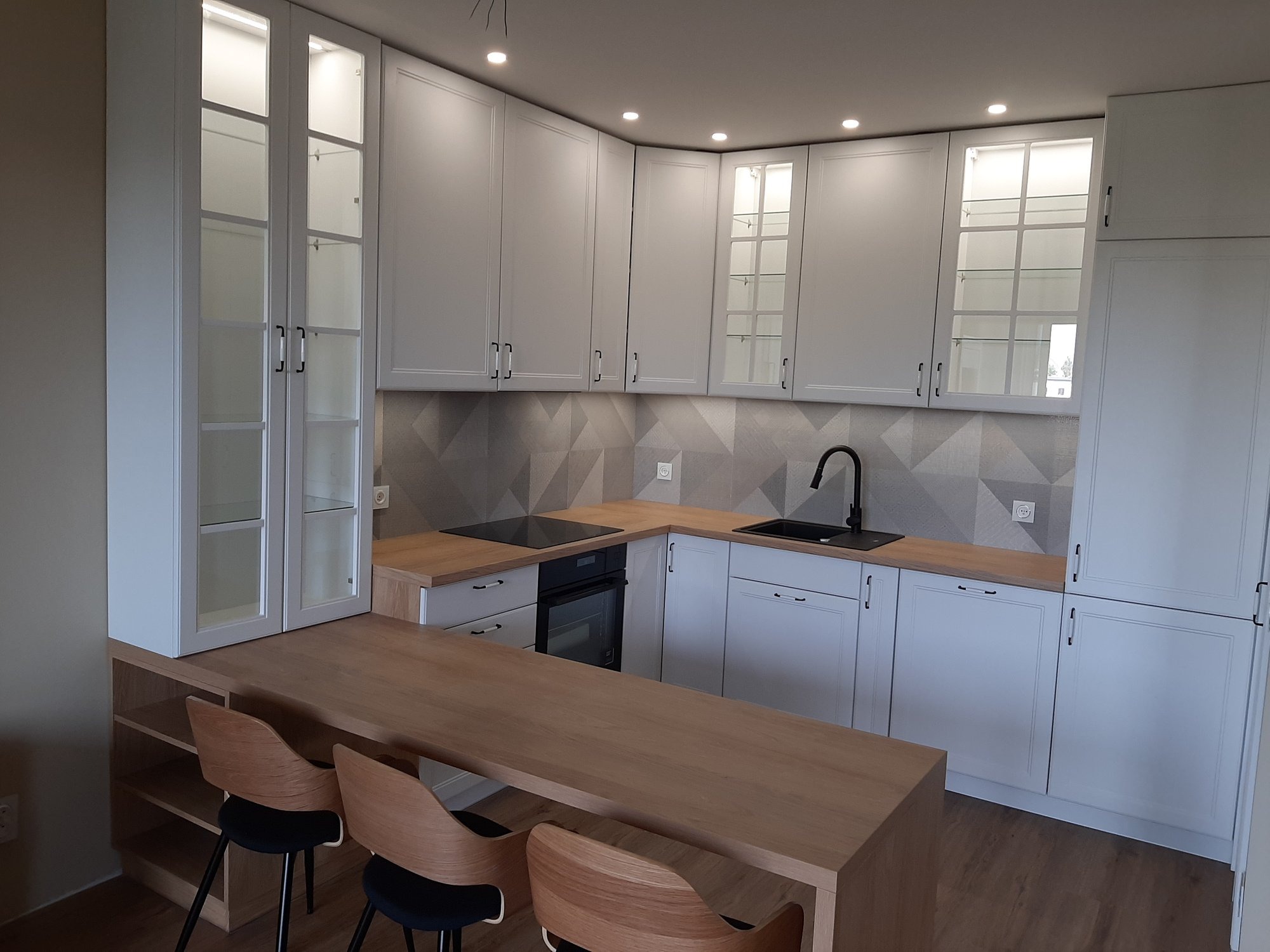 Jasna kuchnia z drewnianym blatem i wyspą, szare płytki na ścianie, przeszklone szafki górne z podświetleniem, nowoczesne krzesła barowe z drewnianym oparciem.