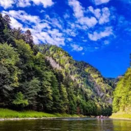 Spływ tratwami po Dunajcu, widok z poziomu rzeki na zalesione zbocza gór i błękitne niebo z chmurami.
