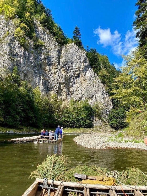 Spływ tratwą po Dunajcu w Pieninach, widok z przodu tratwy na grupę turystów płynących w kierunku skalistego brzegu rzeki otoczonego drzewami.