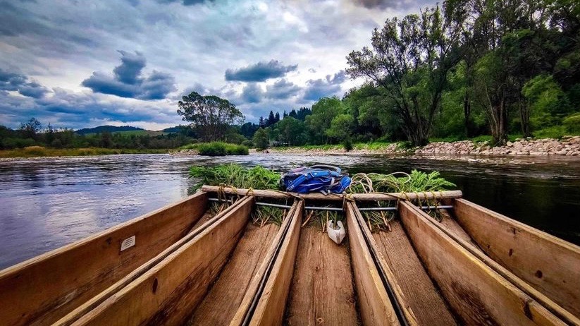 Drewniana tratwa z plecakiem i linami na rzece, widok z perspektywy pasażera, w tle zalesione brzegi i pochmurne niebo.