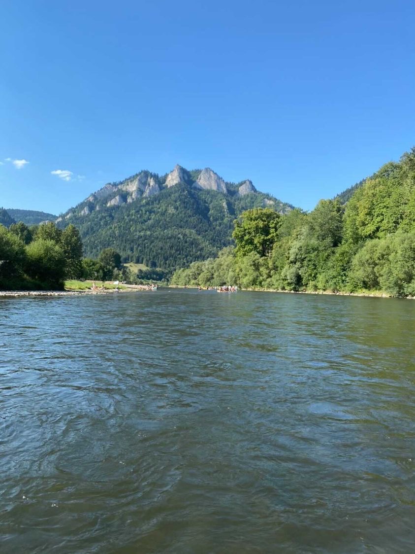 Spokojny nurt rzeki z widokiem na skalisty szczyt góry porośniętej lasem, błękitne niebo w tle, w oddali widoczne pontony z turystami.