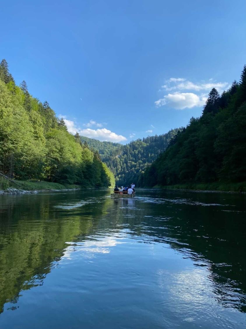 Spokojny spływ tratwą po rzece otoczonej zielonymi, zalesionymi wzgórzami w słoneczny dzień. Woda odbija błękitne niebo.