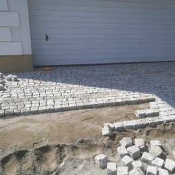 Golden hans - Fragment podjazdu z kostki granitowej w trakcie układania; widoczne przygotowane podłoże, ułożona część nawierzchni oraz kostki gotowe do montażu przy garażu.