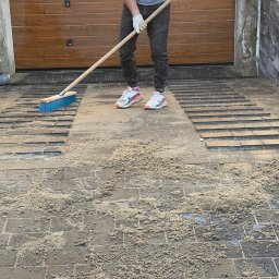 Golden hans - Pracownik w Piasecznie zamiata piasek z ułożonej częściowo kostki brukowej na podjeździe przed garażem; widoczne buty sportowe i białe rękawice robocze.