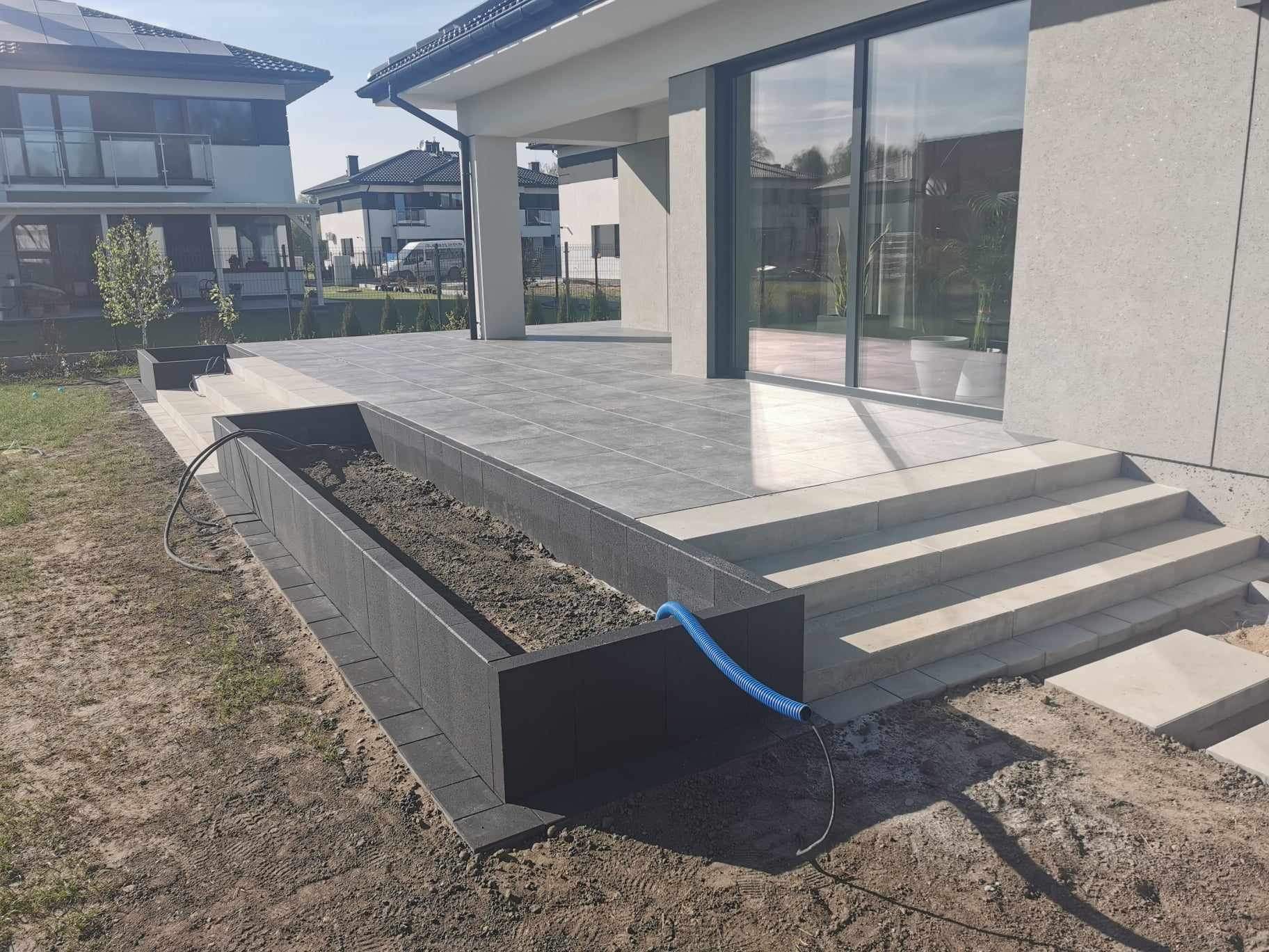 Nowoczesny taras z szarej kostki brukowej, schody prowadzące do domu z dużymi oknami, obok podwyższona rabata z ziemią, z boku leży niebieski wąż ogrodowy.