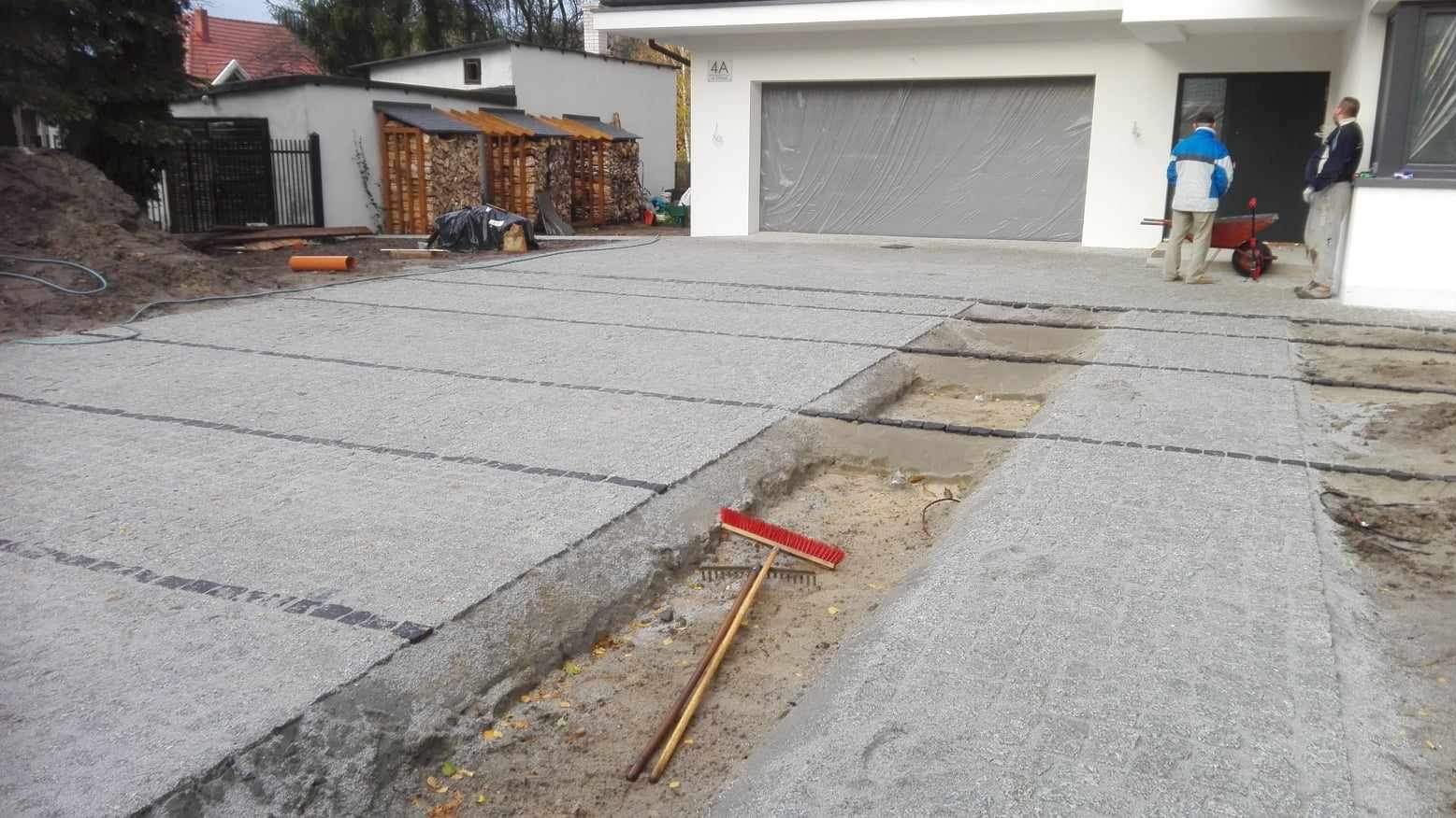 Przygotowanie podłoża pod nawierzchnię z kostki brukowej, widoczne czarne pasy separacyjne, częściowo ułożona kostka wzdłuż krawędzi, czerwona szczotka i grabie leżące na piasku, dwaj pracownicy...