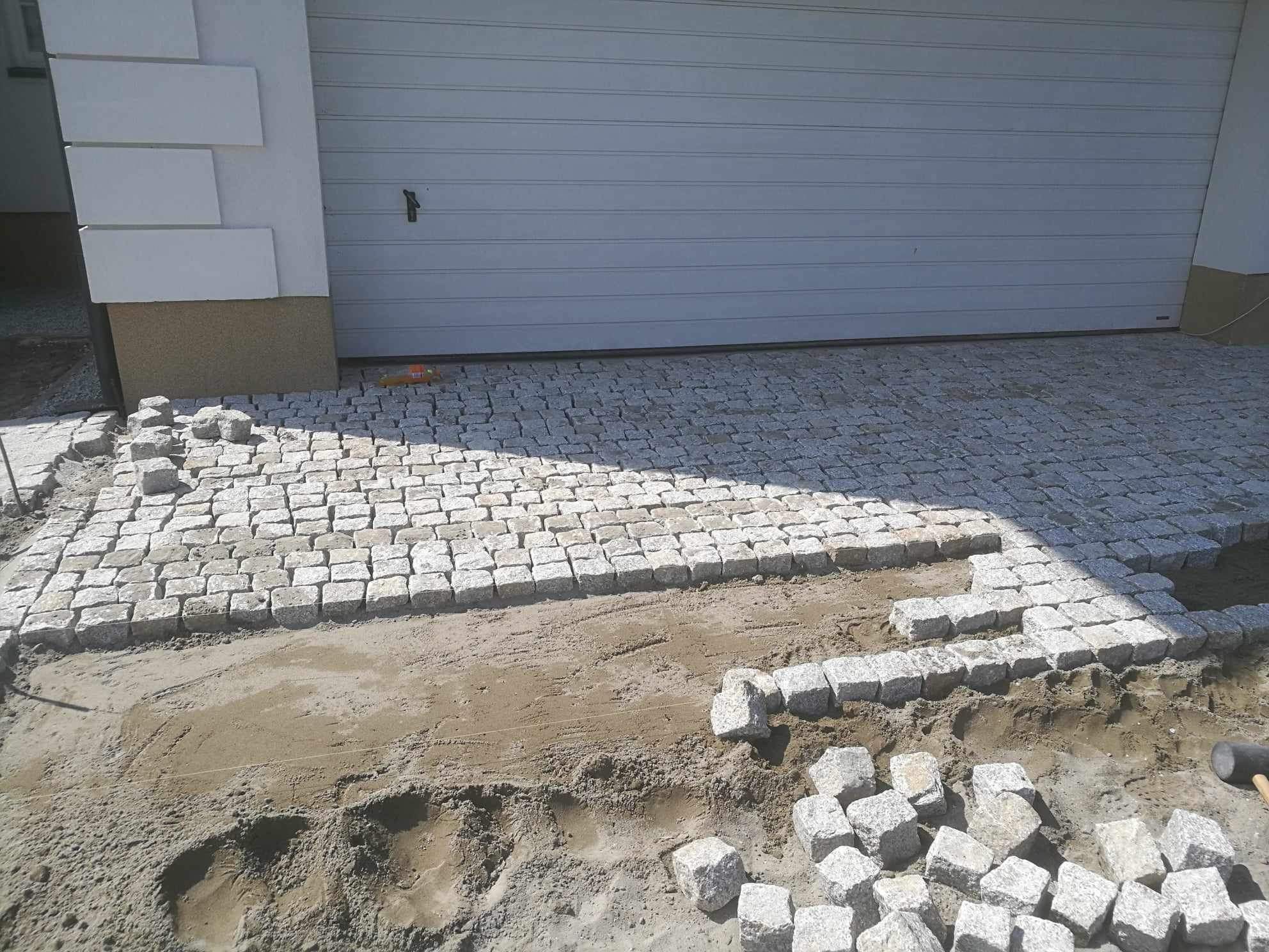 Fragment podjazdu z kostki granitowej w trakcie układania; widoczne przygotowane podłoże, ułożona część nawierzchni oraz kostki gotowe do montażu przy garażu.
