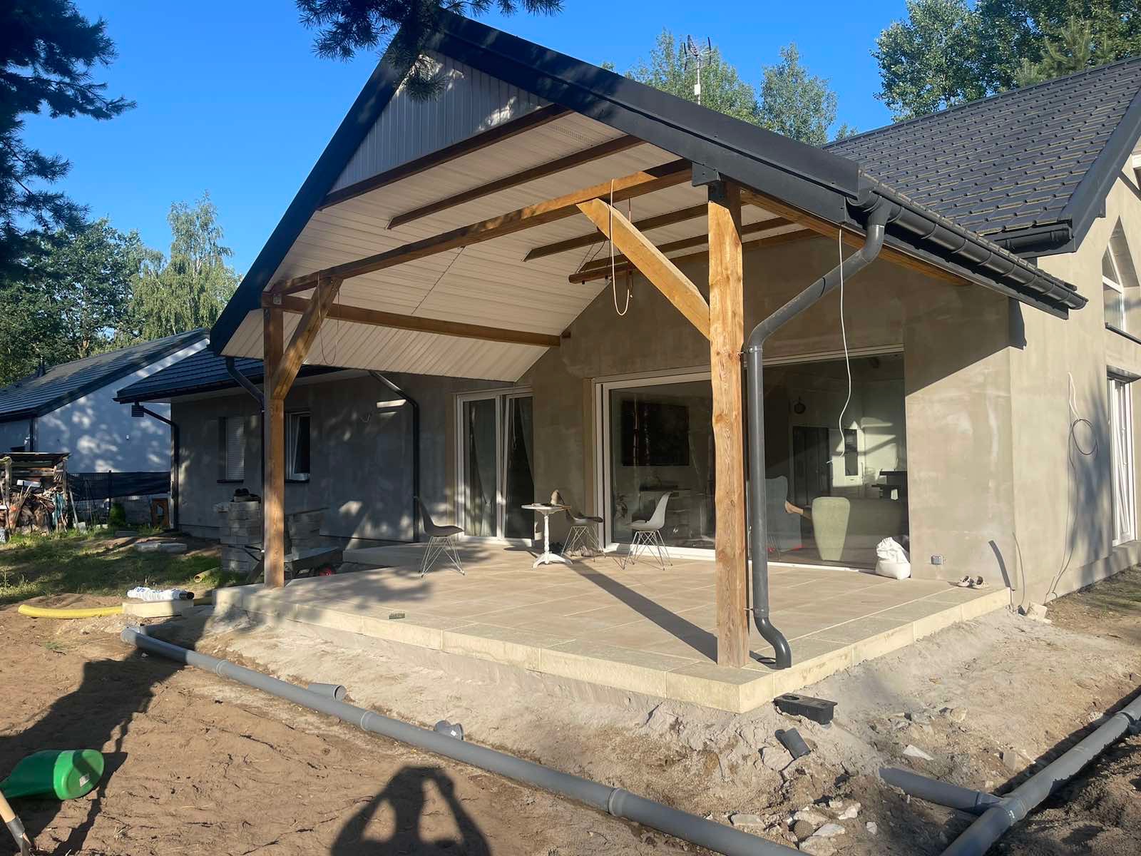 Nowoczesny taras z jasnej kostki, z drewnianą konstrukcją zadaszenia i widocznymi rurami kanalizacyjnymi w gruncie przed tarasem.