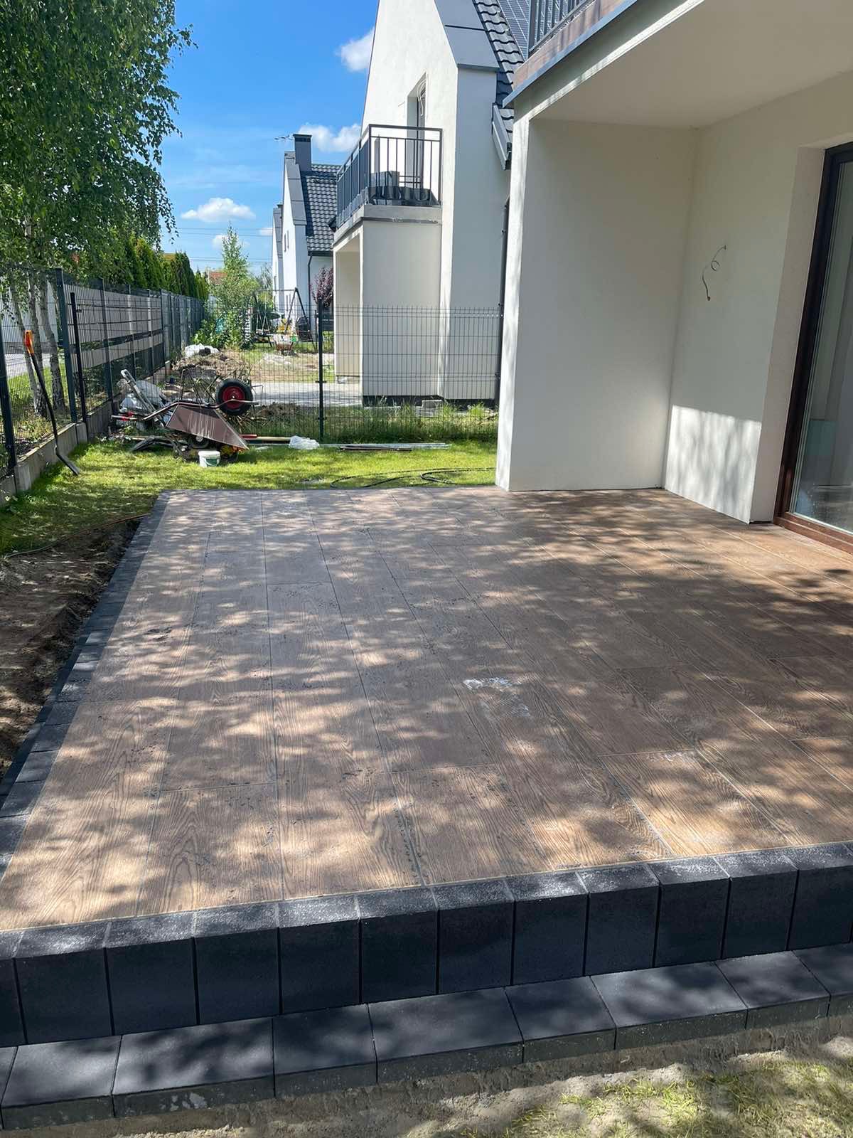 Wykończony taras z desek kompozytowych imitujących drewno, obrzeża z ciemnej kostki brukowej, widok na ogród i fragment elewacji domu.