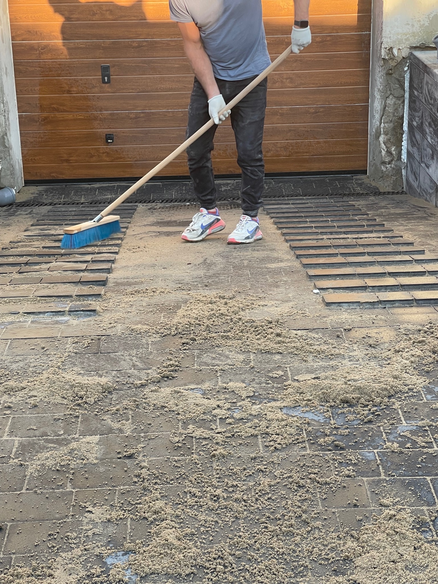 Pracownik w Piasecznie zamiata piasek z ułożonej częściowo kostki brukowej na podjeździe przed garażem; widoczne buty sportowe i białe rękawice robocze.