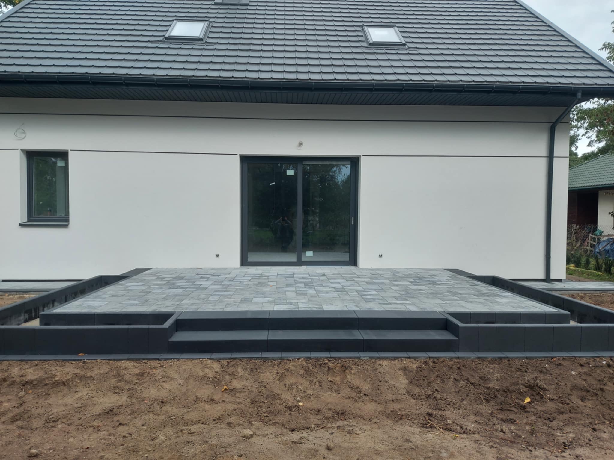 Nowoczesny taras z szarej kostki brukowej z dwustopniowym wejściem, otoczony ciemnoszarą obwódką, na tle białego domu z szarym dachem i oknami.