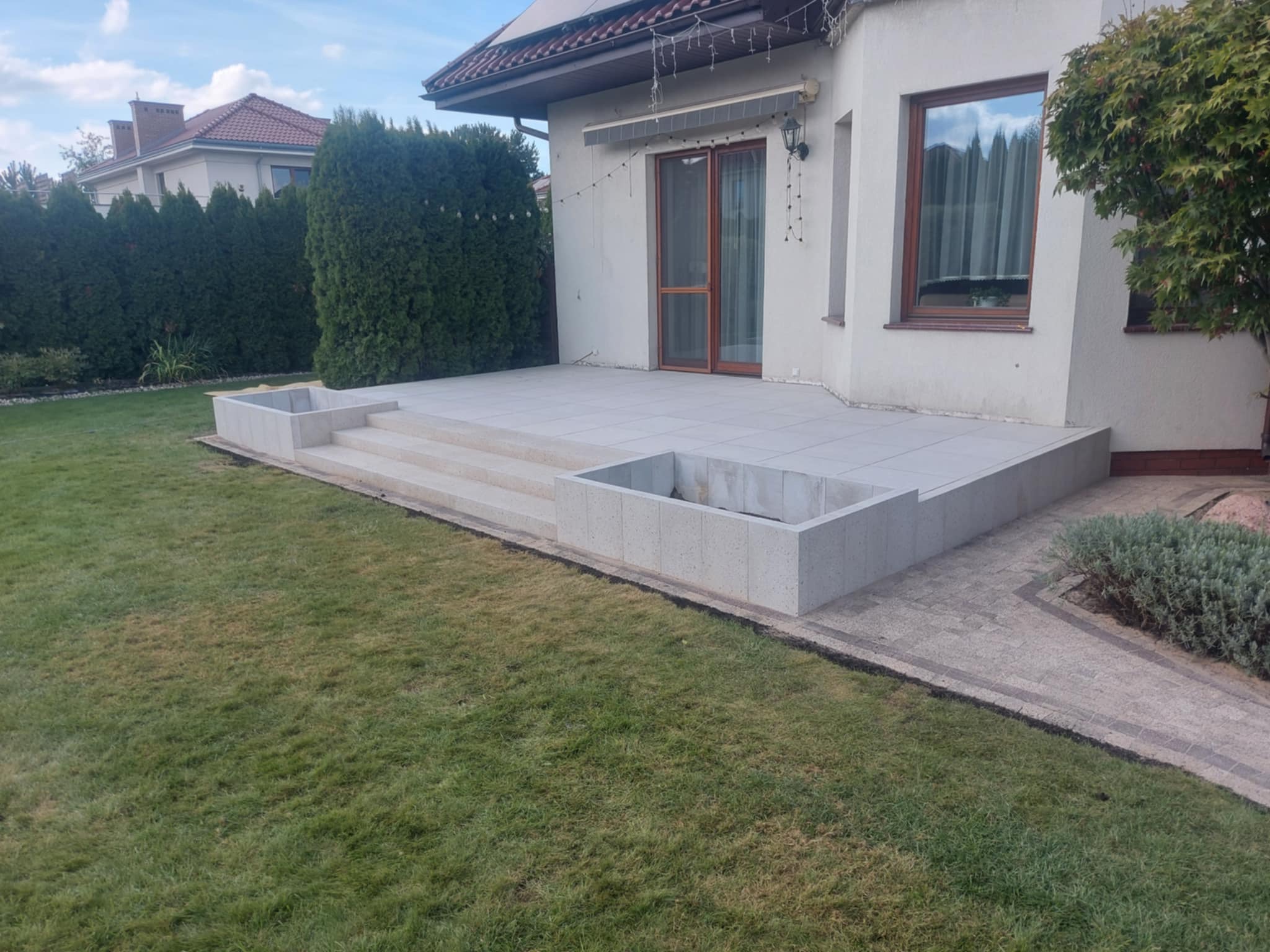 Nowoczesny taras z jasnych płyt, z dwoma podwyższonymi rabatami z betonowych bloczków, schody prowadzące do domu, trawnik i roślinność w tle.