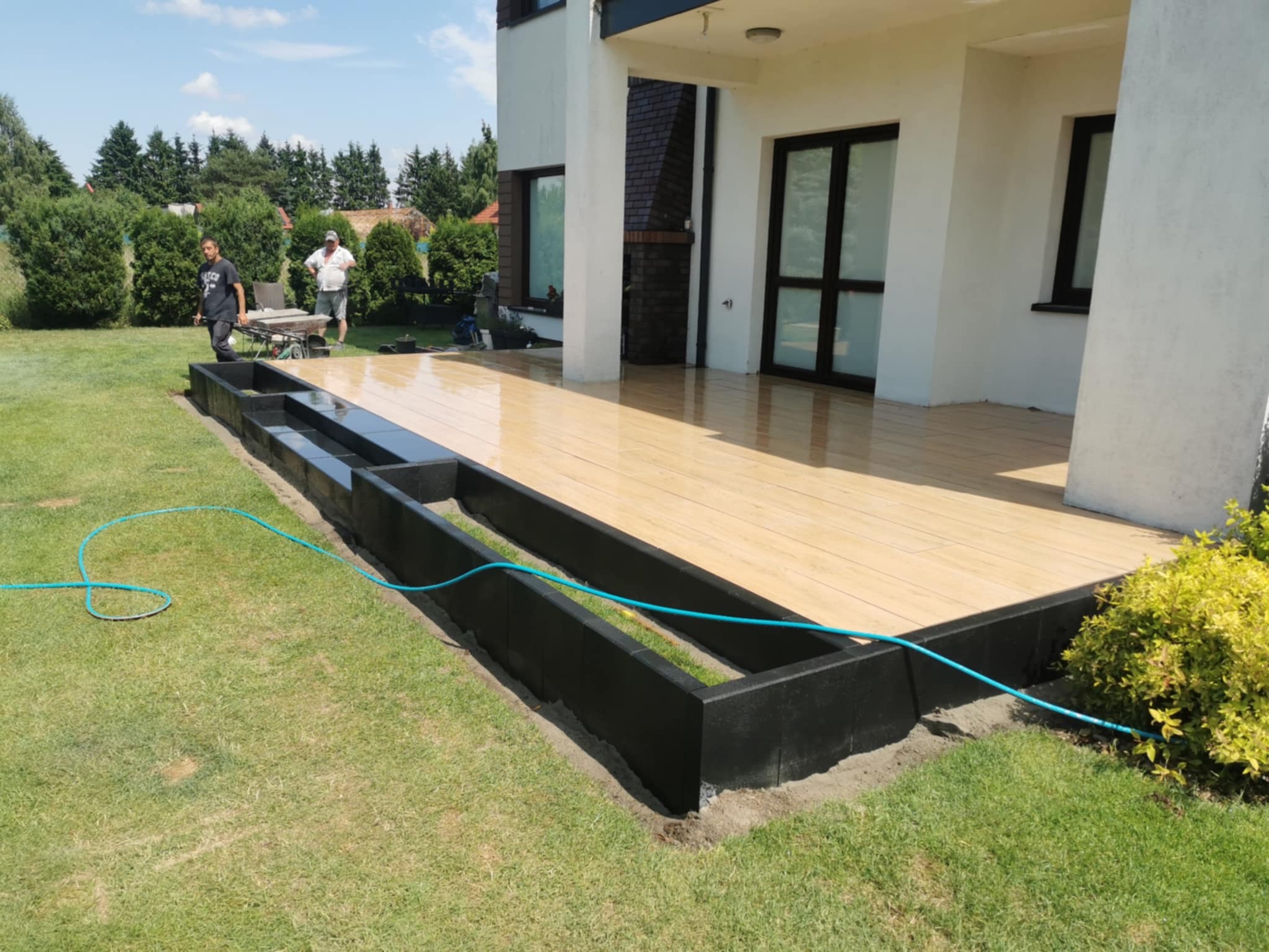 Jasna, drewnopodobna kostka na tarasie domu jednorodzinnego, obok czarne obrzeża rabat. W tle pracownicy i taczka. Błękitny wąż ogrodowy na trawie.