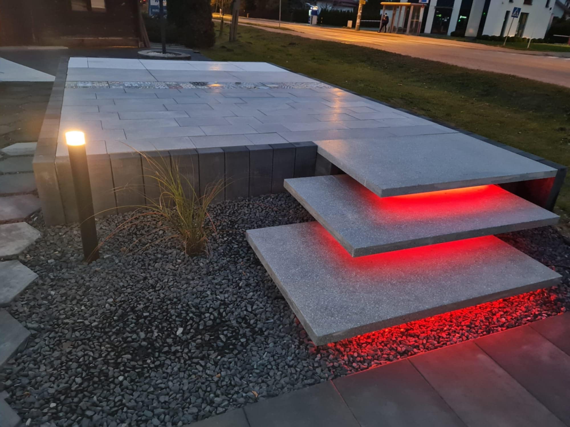 Nowoczesne schody z kostki granitowej z podświetleniem LED, otoczone ozdobnym żwirem i trawą. Widoczne oświetlenie zewnętrzne i fragment chodnika.