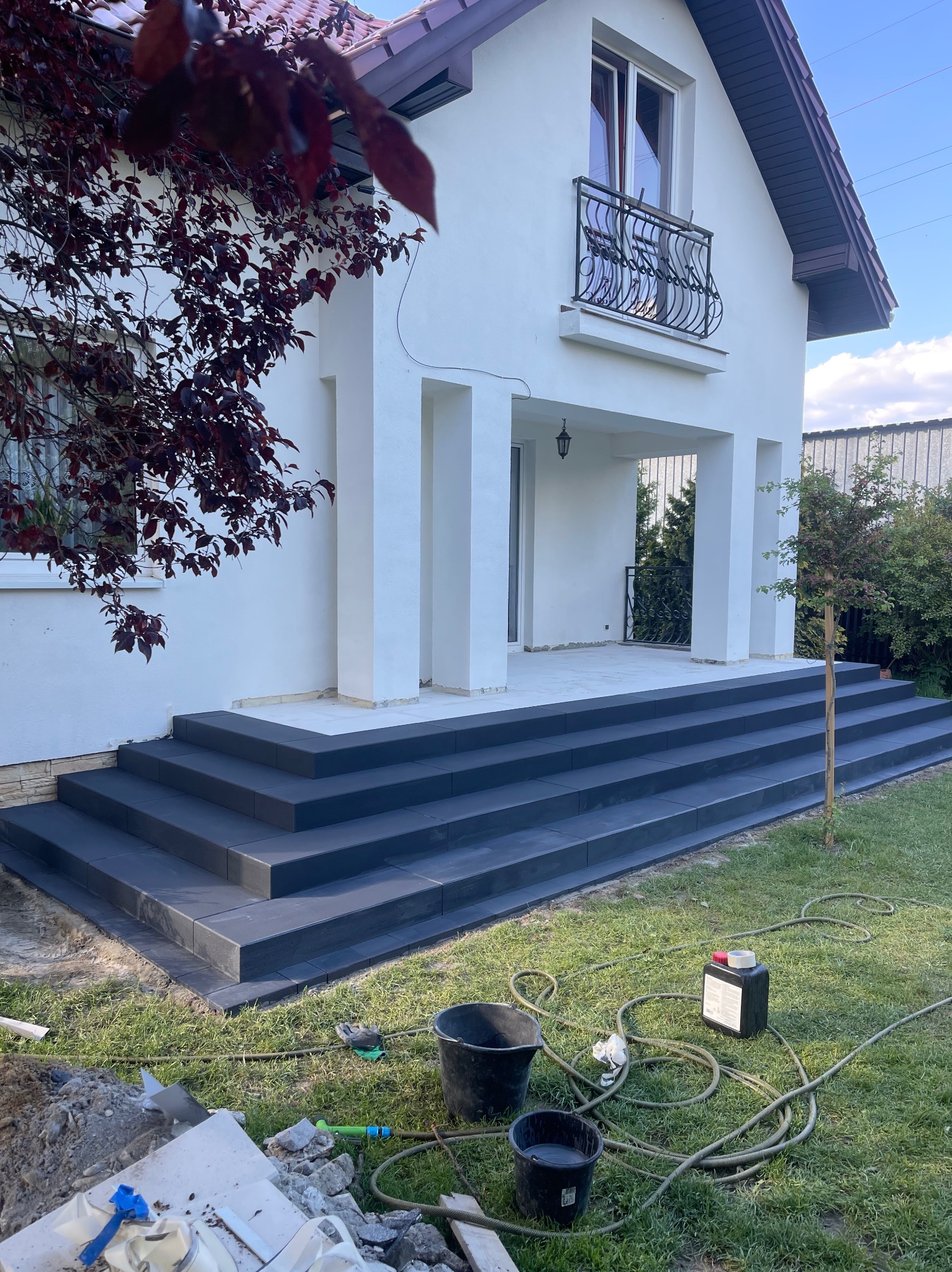 Nowe, ciemne schody z kostki brukowej prowadzące do białego domu z balkonem. Na trawniku widoczne narzędzia i materiały budowlane.