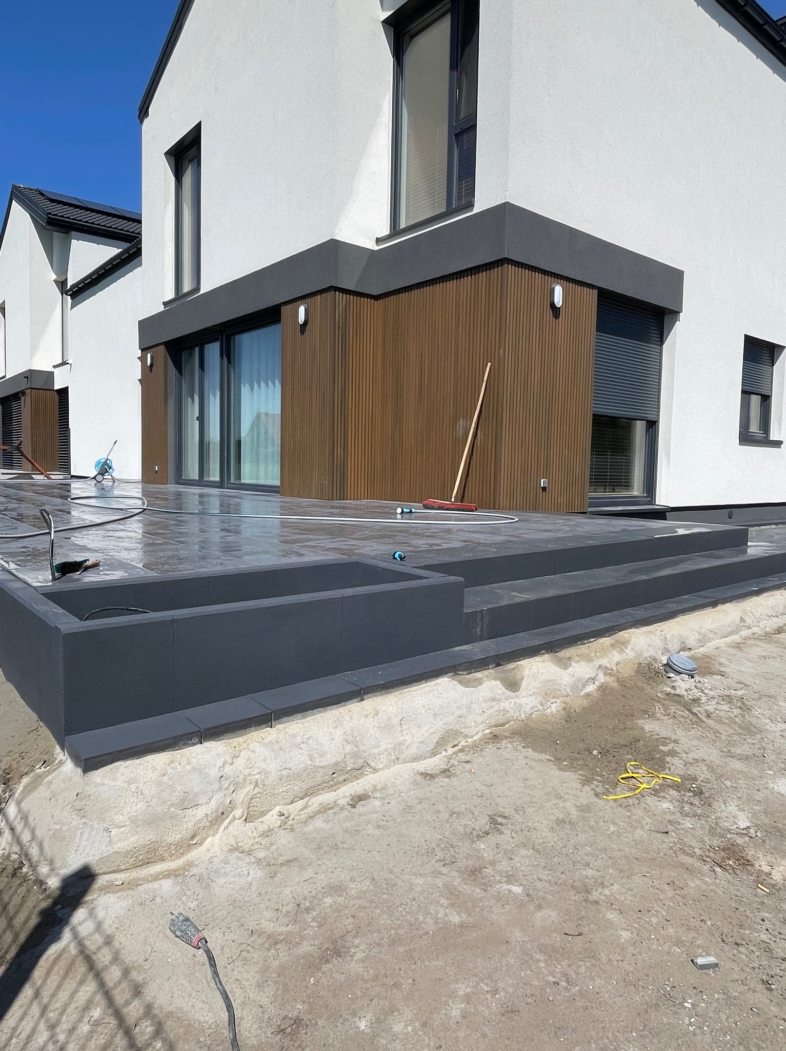 Nowoczesny taras z szarych płyt, częściowo mokry po myciu, z widocznym wężem i szczotką. Tło stanowi biały budynek z elementami drewnianymi i szarą stolarką okienną.