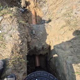 FRYGA AMI Sp. Z o.o. - Wykop z widocznymi rurami kanalizacyjnymi z tworzywa sztucznego i czarną pokrywą studzienki rewizyjnej, obok but roboczy, w tle ziemia i trawa.