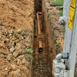 Wykop z ułożonymi rurami kanalizacyjnymi z tworzywa sztucznego, widok z góry na dno wykopu, częściowo zasypane ziemią, z boku widoczny fragment maszyny budowlanej.