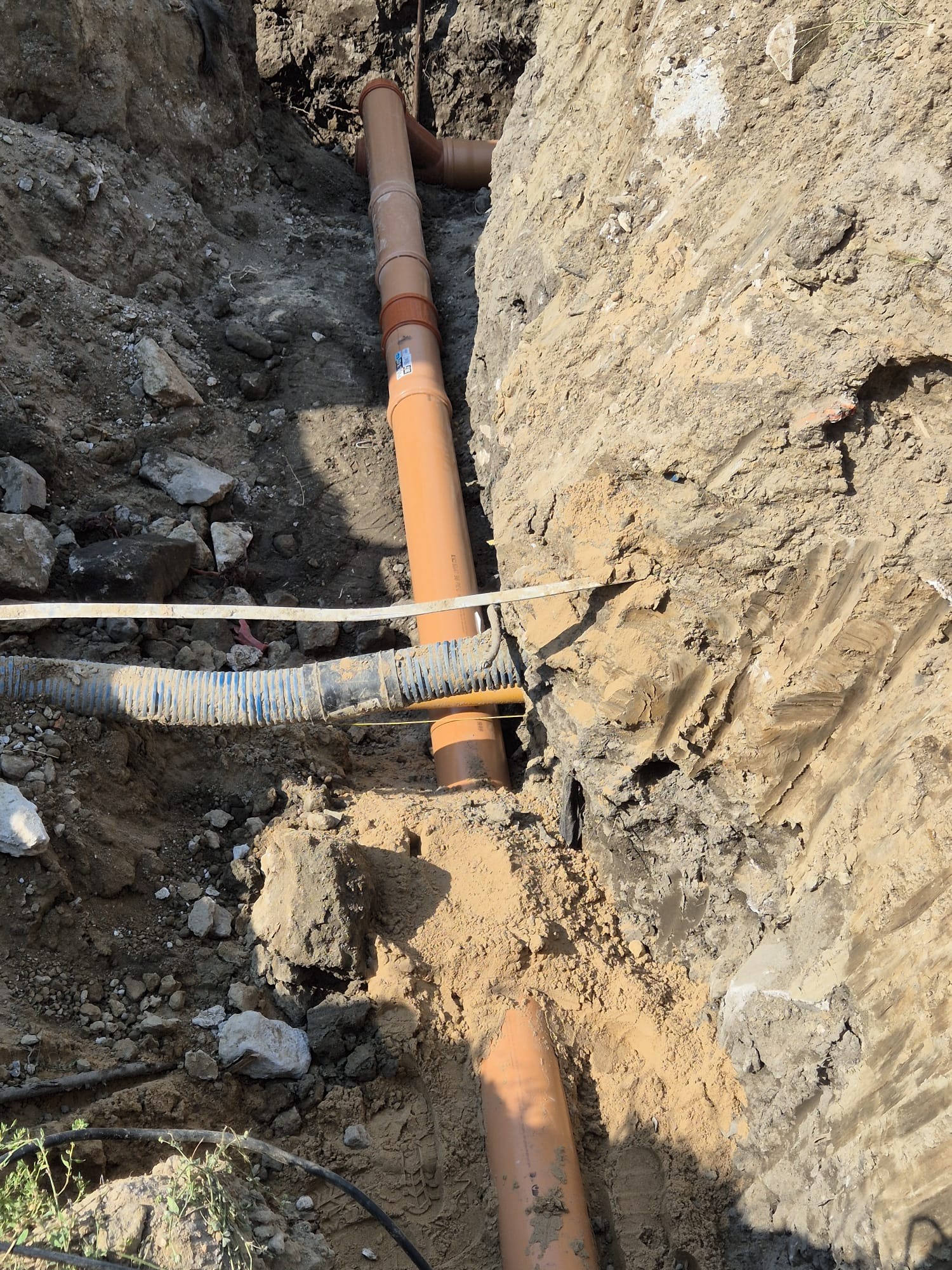 Wykonany wykop pod rury kanalizacyjne z widocznymi elementami przyłącza, w tym rurą karbowaną i pomarańczowymi rurami PVC z uszczelkami, w naturalnym, ziemistym otoczeniu wykopu.