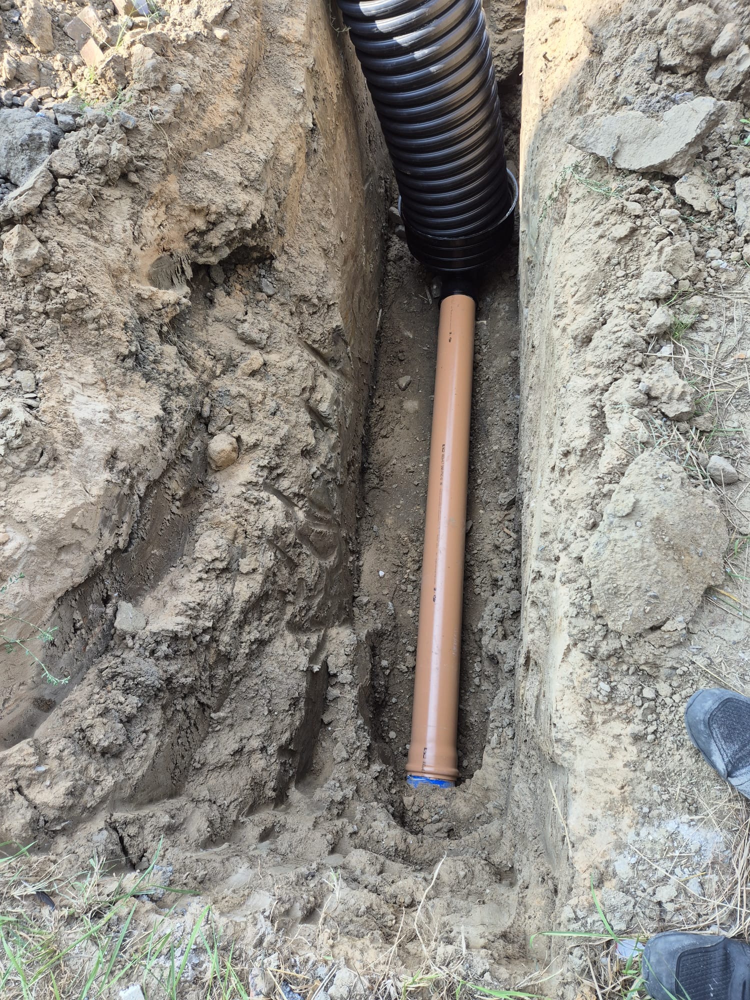 Wykop z ułożonymi rurami: czarną karbowaną i brązową gładką, widoczne ślady pracy ziemnej i fragment buta w rogu.