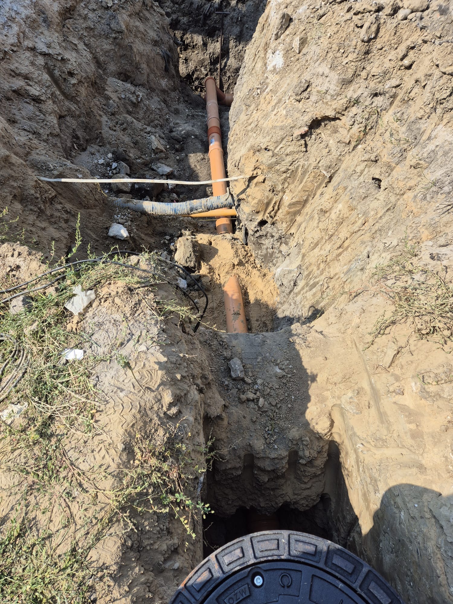 Wykop z pomarańczowymi rurami kanalizacyjnymi w ziemi, widoczne złącza i karbowana rura, zarośnięte trawą brzegi wykopu, studzienka kanalizacyjna w dolnej części kadru.